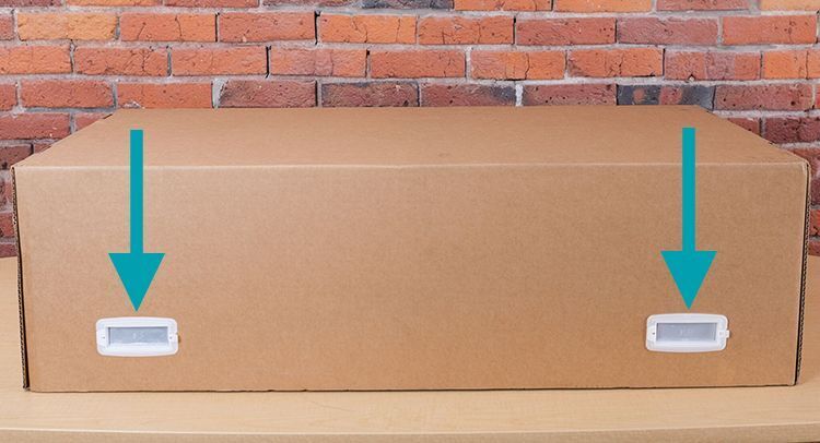 The box a Glowforge arrives in, with arrows pointing to the white plastic handles near the corners of the box.