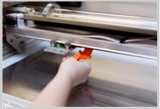 A hand securing an orange platic clip to the laser arm and crumbtray inside a Glowforge