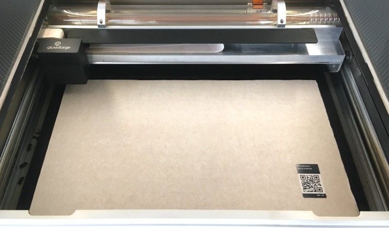 A piece of Glowforge Medium Draftboard placed in the bed of a Glowforge printer with the lid open